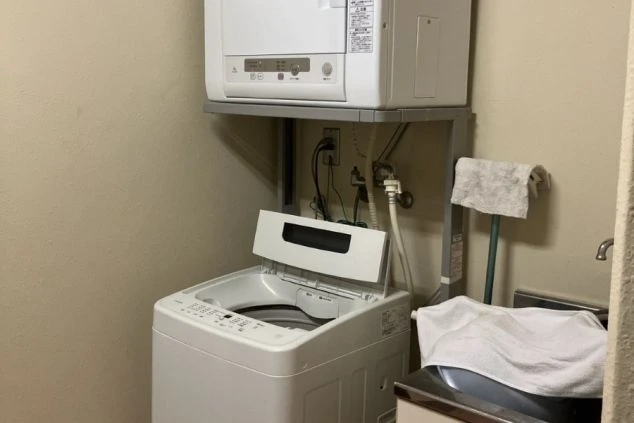 寮の共用ランドリースペース。縦型の全自動洗濯機の上に、専用のラックで衣類乾燥機が設置されている。限られたスペースを有効活用した清潔感のある設備。