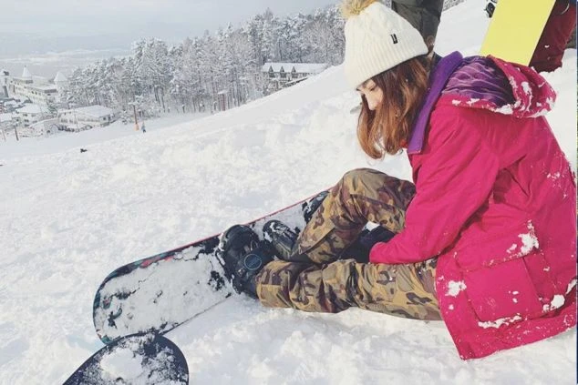雪山でスノーボードの準備をする女性。白いニット帽とピンクのウェアを着用し、奥にはスキー場の建物が見える。