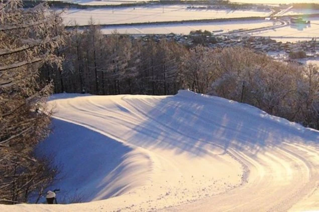 日が当たり、雪面が美しく波打つ丘。遠くには雪景色のまちが見える。