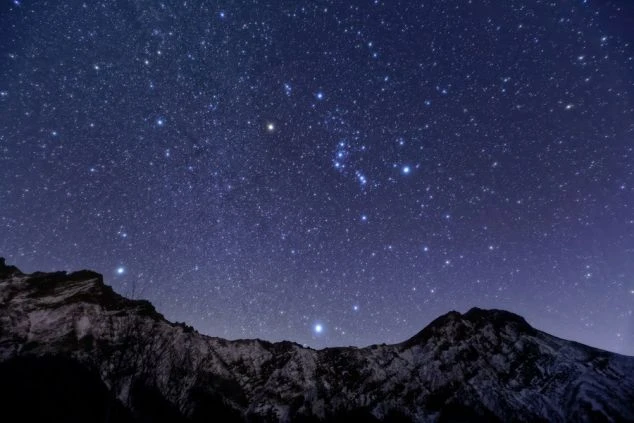  暗い山々の稜線の上に広がる満天の星空。無数の星が輝き、オリオン座もはっきりと見える。