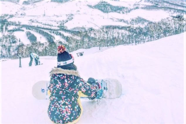 雪山の斜面でスノーボードをする人物の後ろ姿の画像。カラフルなウェアを着用し、景色を眺めている。リゾートバイトの休日にウィンタースポーツを楽しめる。