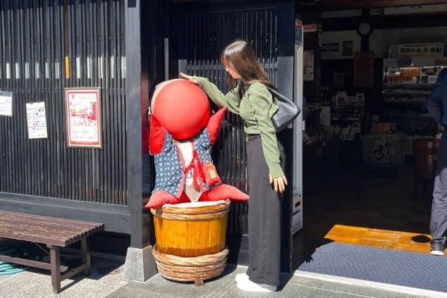 ワクトリ社員が撮影した、伝統的な建物の前で、赤い「さるぼぼ」の置物に触れる女性。