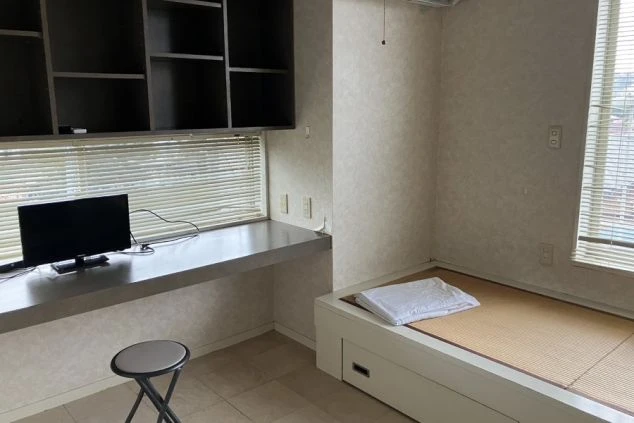 寮の個室。布団が敷かれた台と、テレビ付きの長い机、棚、窓にブラインドが映る。