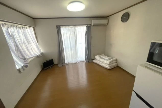 寮の個室。布団とテレビ、エアコン、時計、窓にカーテンが映る部屋の様子。