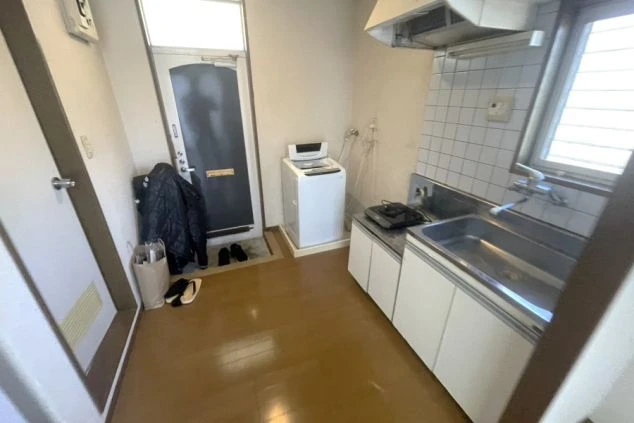 寮の玄関。ドアの横に洗濯機、キッチン、シンク、コンロが並ぶ生活空間が映る。