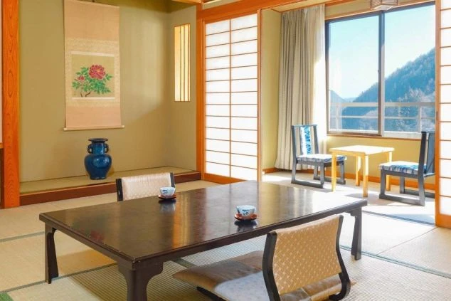 和室の客室の画像。畳敷きの空間に座卓と座椅子が置かれ、窓からは明るい日差しと山の景色が見える。リゾートバイト中の滞在環境をイメージ。
