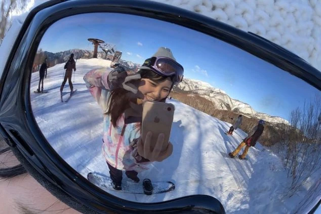 スキー場で自撮りをする女性のゴーグルに映る景色。笑顔の女性と雪山、スキーヤーが見える。