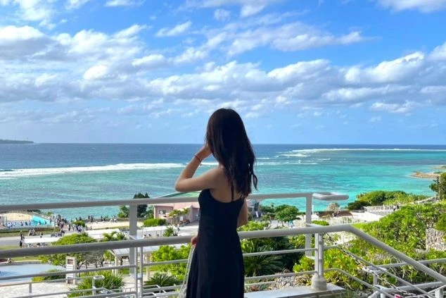ホテルのバルコニーから、エメラルドグリーンに輝く海を眺める女性。沖縄の美しい景色が広がる。