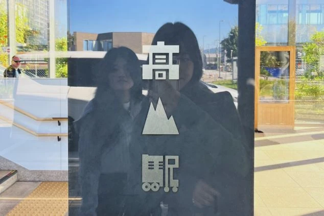 飛騨高山駅の入口付近のガラスに映る二人の人物の姿。ガラスには「高山」の文字と山のイラストが描かれ、駅周辺でのリゾートバイトや、観光地へのアクセスに興味がある方に訴求するワクトリオリジナル画像。