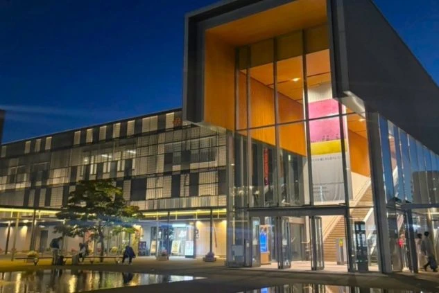 夜のJR高山駅のモダンな駅舎。ガラス張りの建物がライトアップされ、駅前広場に光が反射している。交通の便が良く、仕事後のアクセスも便利なリゾートバイトの勤務地をイメージさせる画像。