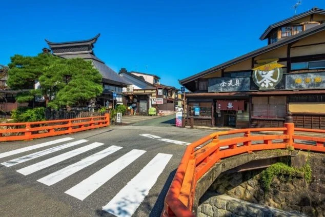 青空の下、赤い橋と趣のある建物が並ぶ飛騨高山の街角。観光客が行き交う賑やかな風景で、リゾートバイトでお土産物店や飲食店での接客に興味がある方に訴求する画像。