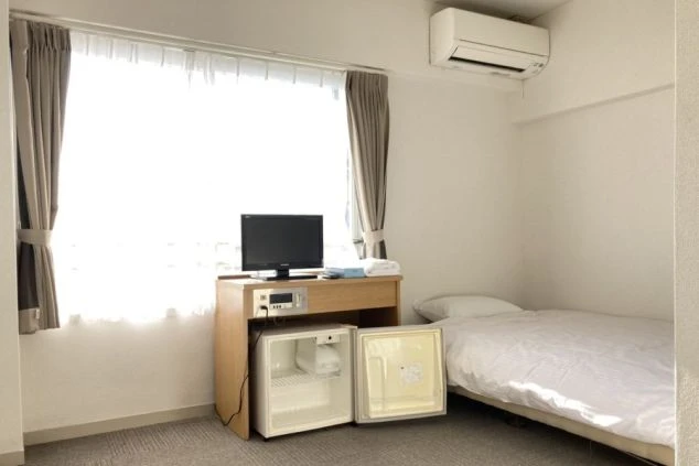 リゾートバイトの個室寮の室内。シングルベッドの横にテレビとミニ冷蔵庫が置かれ、窓からは明るい光が差し込む、快適に過ごせる寮の部屋の画像。