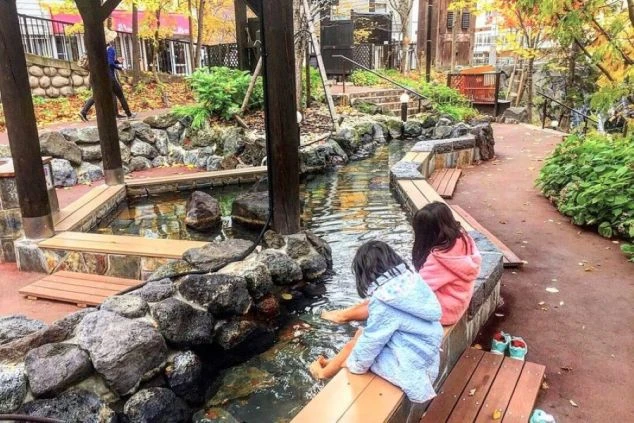 紅葉の季節の足湯。2人の子供が湯船に足を入れており、石や木が配された和風の空間。