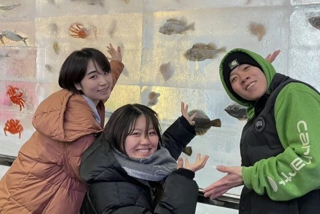  氷の壁に魚が埋め込まれた壁で、笑顔でポーズをとる3人の若者たち。冬のリゾートバイトの思い出。