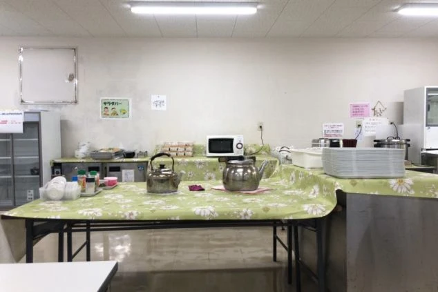 リゾートバイト寮の広々とした食堂。花柄のテーブルクロスが敷かれたテーブルと、電子レンジや給湯器が設置されたセルフサービスエリアがある、従業員食堂の画像。