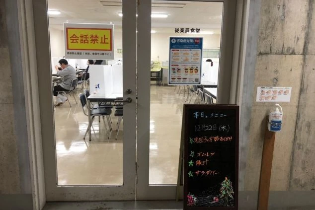 「従業員食堂」と書かれたドア越しに、仕切りが設置されたテーブルで食事ができる従業員食堂。本日のメニューが書かれた黒板も見える、感染症対策がされている食堂の画像。