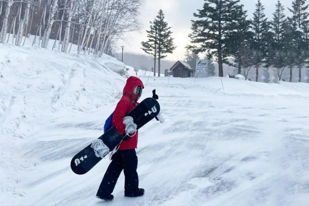 雪景色のスキー場でスノーボードを担いで歩く人物の後ろ姿。リゾートバイトでスキー場スタッフとして働きながら、ウィンタースポーツを楽しみたい方に訴求する画像。