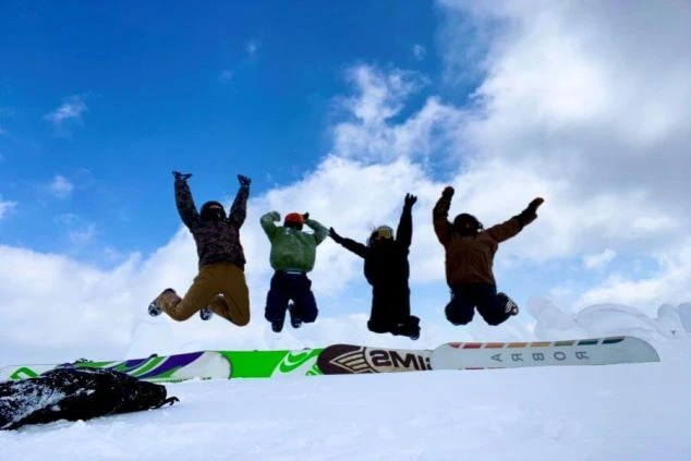 青空の下、雪山でスノーボードの板を前に並んでジャンプする4人のスノーボーダーのシルエット。リゾートバイトで出会った仲間と、冬のゲレンデで最高の思い出を作れることを示すワクトリオリジナル画像。