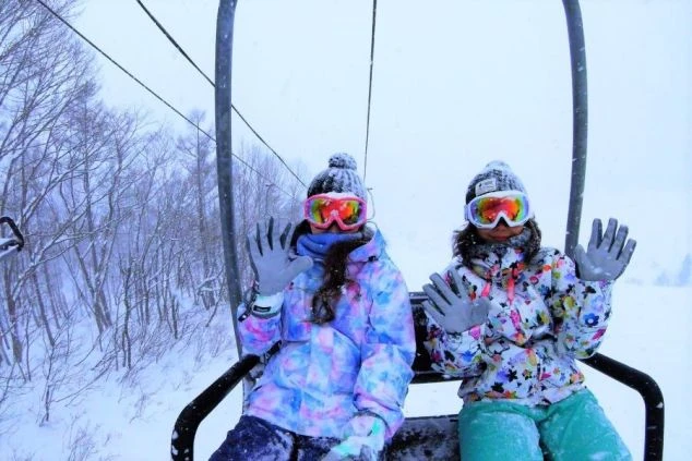 雪が降る中でスキーリフトに乗って手を振るカラフルなスキーウェアとゴーグルを着用した2人の女性。冬のリゾートバイトでスキー場での仕事や、雪山でのアクティビティを楽しみたい方に魅力的なスタッフ画像。