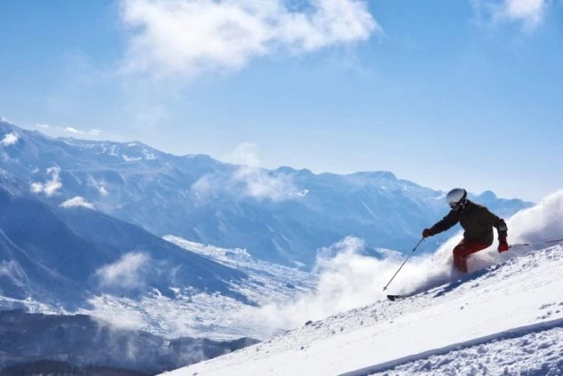 青い空の下、雪山を豪快にスキーで滑り降りる人物。遠くには雪化粧した山々が連なる絶景。