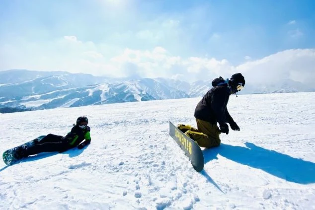 雪山のゲレンデでスノーボードを楽しむ2人の人物。転んだり、起き上がったりしながら、ウィンタースポーツを満喫している。リゾートバイトの休日の過ごし方のイメージ。