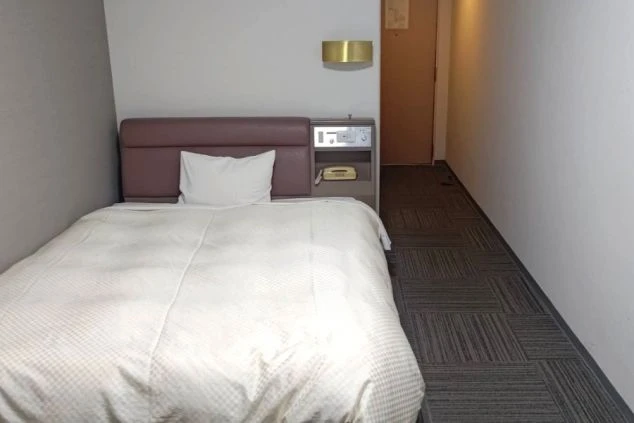 リゾートバイトの寮の部屋の画像。枕元の壁に照明が取り付けられ、ベッドと電話が置かれている。リゾートバイトの寮の部屋の画像。