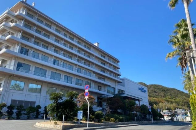 ヤシの木が植えられた道路沿いに建つ、白いホテルや旅館の外観の画像。ヤシの木と青空が南国リゾートのような雰囲気を出している。リゾートバイトの仕事場となる、ホテルの外観の画像。