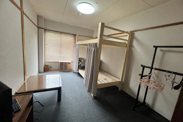 リゾートバイトの寮の部屋の画像。二段ベッドとテーブル、テレビが置かれ、窓にはブラインドがある。プライベート空間が確保された居住空間。