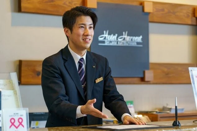 ホテルのフロントで笑顔で顧客対応をする男性スタッフ。ネームプレートが見え、ホテルのフロント業務や接客に興味があるリゾートバイト希望者に魅力的な画像。