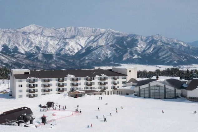 雪山を背景に広がるスキー場とホテル。多くのスキーヤーやスノーボーダーが滑走しており、冬のリゾートバイトでスキー場やホテルスタッフとして働きたい方に魅力的な画像。