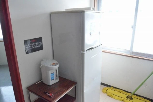 リゾートバイト寮の共用冷蔵庫と電気ポット。窓から明るい光が差し込み、自炊や飲み物の保存に便利な設備の画像。