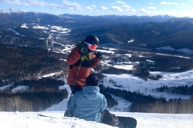 雪山の上でスキーとスノーボードを楽しむ2人の人物。広大な雪景色とリゾート施設を背景に、冬のリゾートバイトでウィンタースポーツを満喫できることを示すワクトリオリジナル画像。