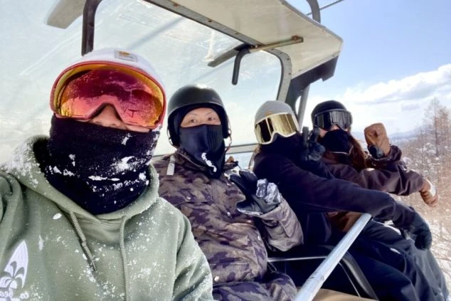 スキーリフトに乗って笑顔でカメラ目線を送る4人のスノーボーダー。雪が降る中で、冬のリゾートバイトで出会った仲間とスキー場での楽しい時間を過ごしていることを示すワクトリオリジナル画像。