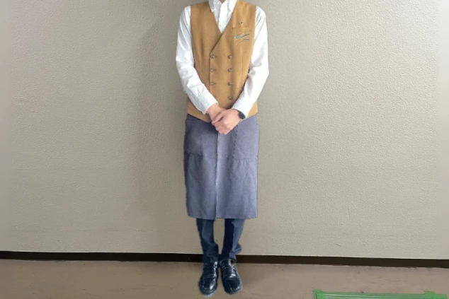 白い長袖シャツに茶色のダブルボタンベスト、濃紺のパンツを着用し、グレーのエプロンを重ねた男性料飲部スタッフの全身画像。