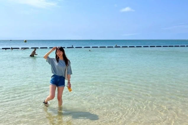 沖縄の透き通った海で、浅瀬に立ってポーズをとる女性。足元までくっきりと見える、美しいエメラルドグリーンの海を満喫している。