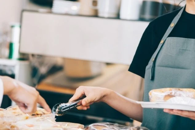 エプロンをつけたスタッフが、お客様の選んだパンをトングを使って取り上げている様子。ベーカリー販売のリゾートバイトの業務イメージ。