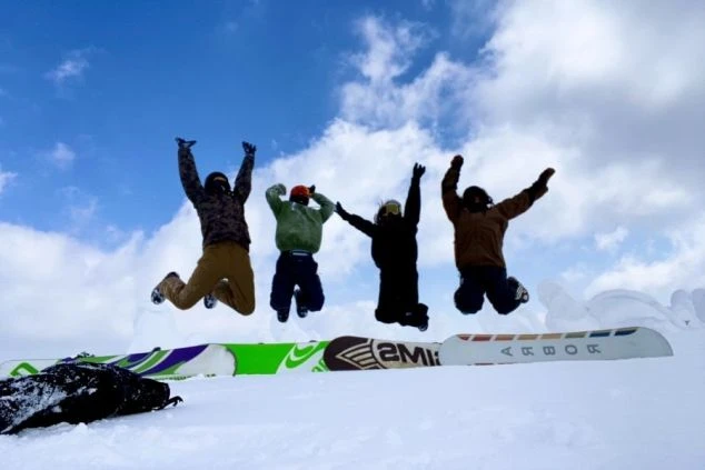 スキー場の山頂で、スノーボーダーのグループが、青空に向かって一斉にジャンプしている。仲間との楽しい思い出の一枚。