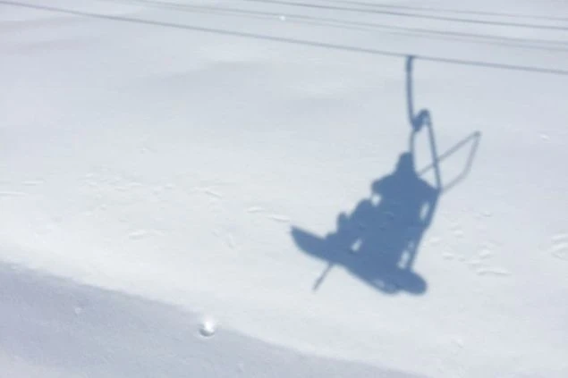 スキー場のリフトに乗っているスノーボーダーの影が、真っ白な雪面に映っている、静かで穏やかな冬の一場面。