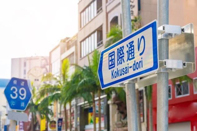 沖縄・那覇のメインストリート「国際通り」の道路標識。背景には、ヤシの木と建物が並ぶ通りの様子が写っている。