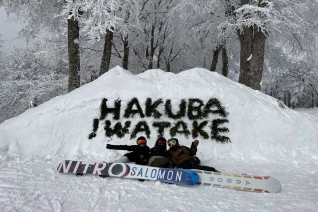 雪山のゲレンデに積もった雪で、「HAKUBA IWATAKE」と書かれた巨大な雪のオブジェの前で、スノーボードを持った3人が座ってポーズを取っている画像です