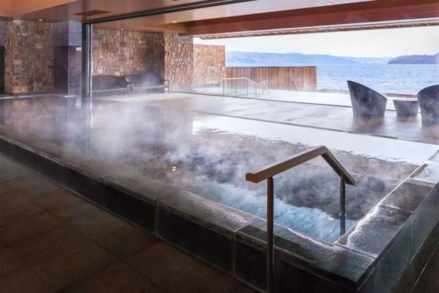 湯気が立ち上る内湯と、その奥の窓が開放されて一体となった露天風呂。浴槽の向こうには湖や海といった水辺の景色が広がり、リラックスチェアが置かれている。
