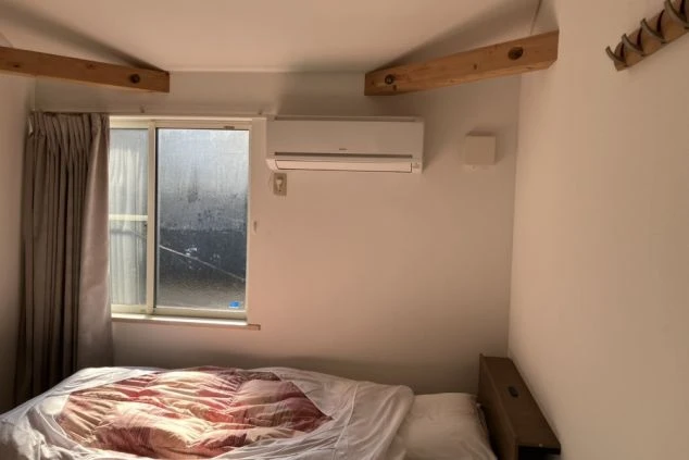 寮の居室。シングルベッドが置かれ、窓の上にエアコン、天井には木製の梁が見える。