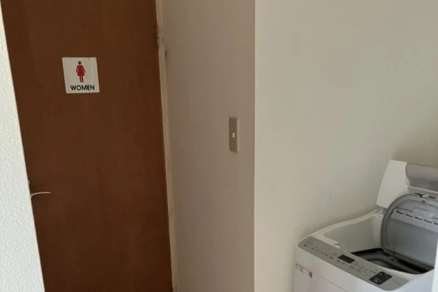 寮の共用エリア。全自動洗濯機の隣に、女性のピクトグラムと「WOMEN」の文字があるトイレまたは更衣室のドアが見える。