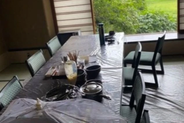 寮の共用食堂または休憩室。窓際に置かれたテーブルの上に食事の準備があり、窓の外には緑の景色が広がっている。和室にテーブルと椅子が置かれた空間。