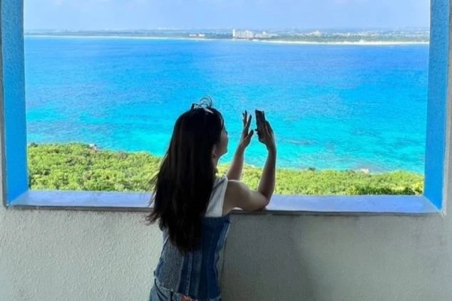 窓から広がる絶景をスマートフォンで撮影する女性。エメラルドグリーンに輝く海が印象的。