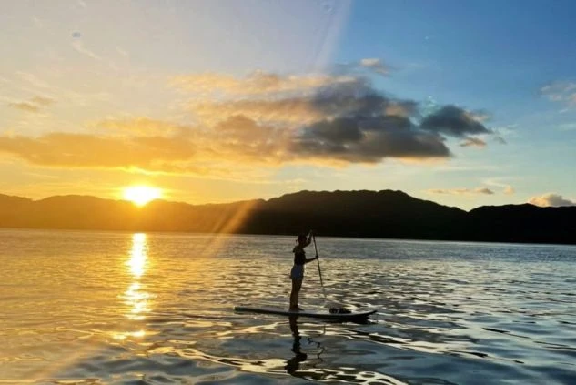 夕焼けの海でSUP（スタンドアップパドルボード）に乗る人物。水面に反射する夕日が幻想的な雰囲気を醸し出す。