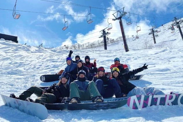 雪山のゲレンデで、スノーボードを持った若者たちが集合写真を撮っている画像。リゾートバイトの仲間との絆が魅力。