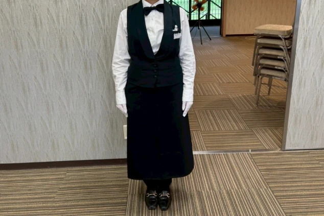 ホールスタッフ(女性)制服。白いブラウスに黒のベストと蝶ネクタイのエレガントなスタイル。レストランサービスのリゾートバイト。