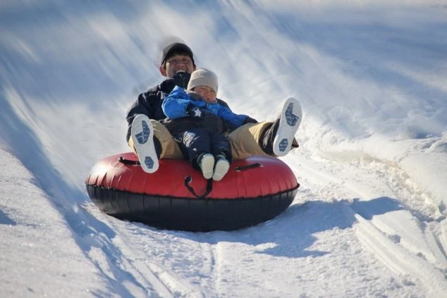リゾートバイトの勤務先スキー場のキッズパークで父親と子どもが二人乗り用の大きな浮き輪（チューブ）に乗って、雪の斜面を滑り降りている様子。