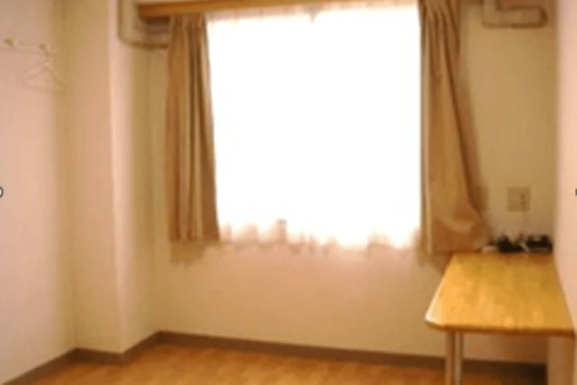 寮の個室。フローリングの床に窓、カーテン、机、ハンガーが映る部屋の様子。